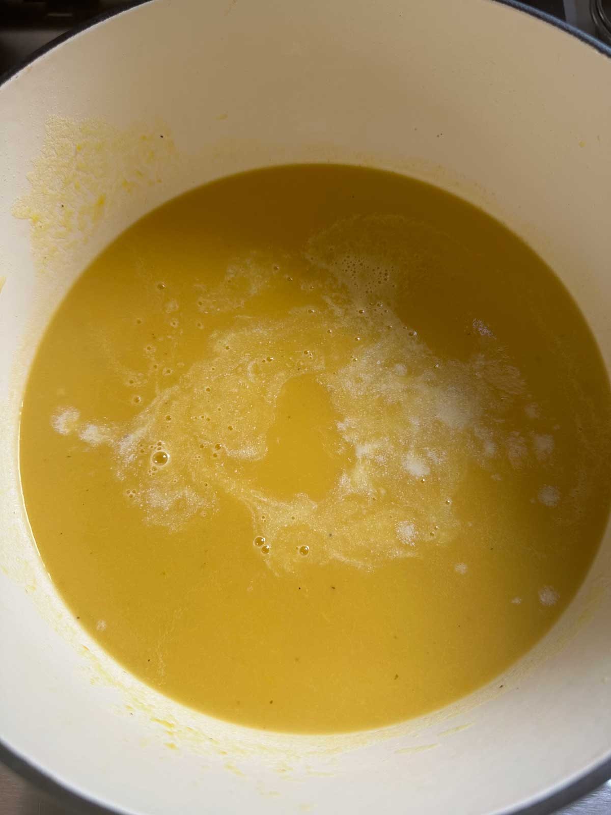 A pot filled with smooth, yellow-orange soup, possibly pumpkin or squash, with small bubbles and a light frothy layer on the surface.
