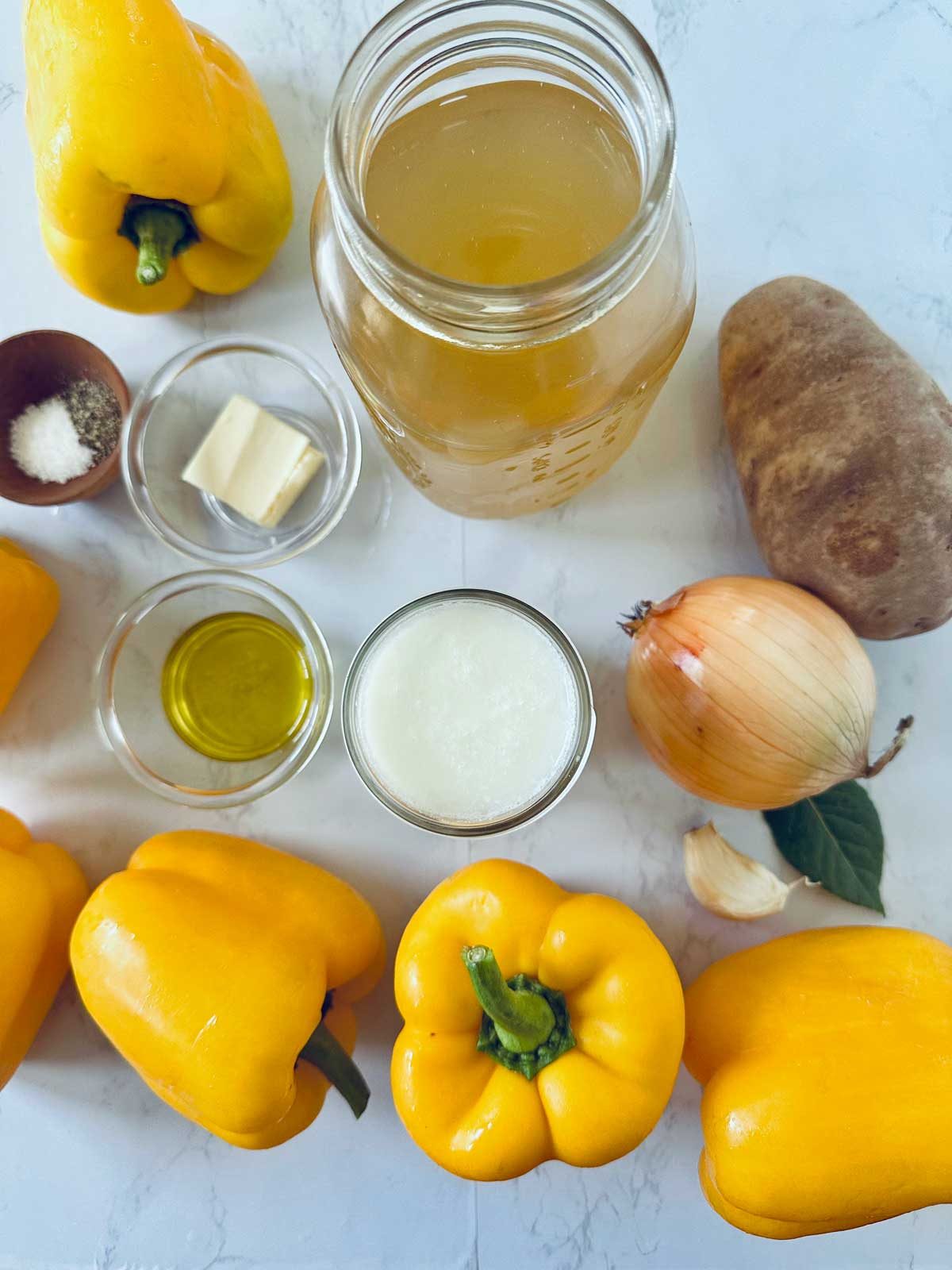 Overhead view of raw ingredients on a white surface: yellow bell peppers, potato, onion, garlic, a jar of broth, a glass of cream, olive oil, butter, salt, and pepper.