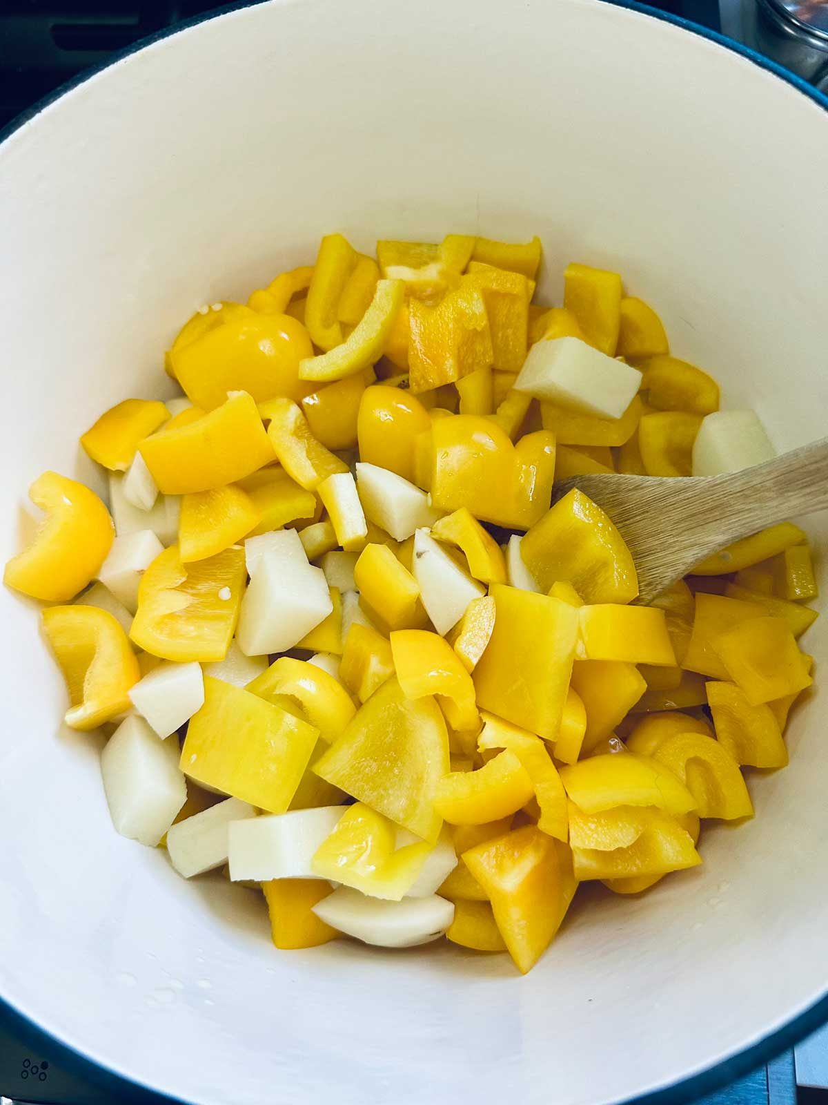 A white pot contains chopped yellow bell peppers and onions being stirred with a wooden spoon.