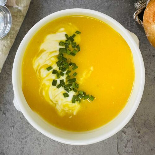 A bowl of creamy yellow soup topped with a swirl of cream and chopped chives, placed on a gray surface next to a piece of bread.