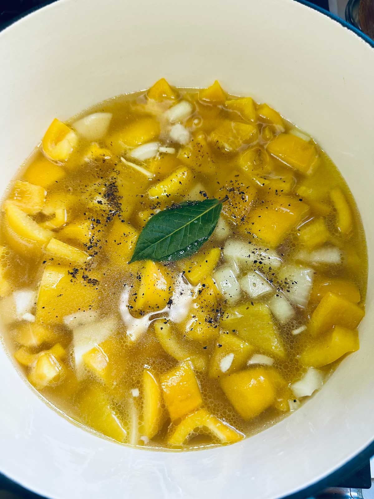 A pot filled with diced yellow squash, chopped onions, broth, a bay leaf, and cracked black pepper, ready to cook.