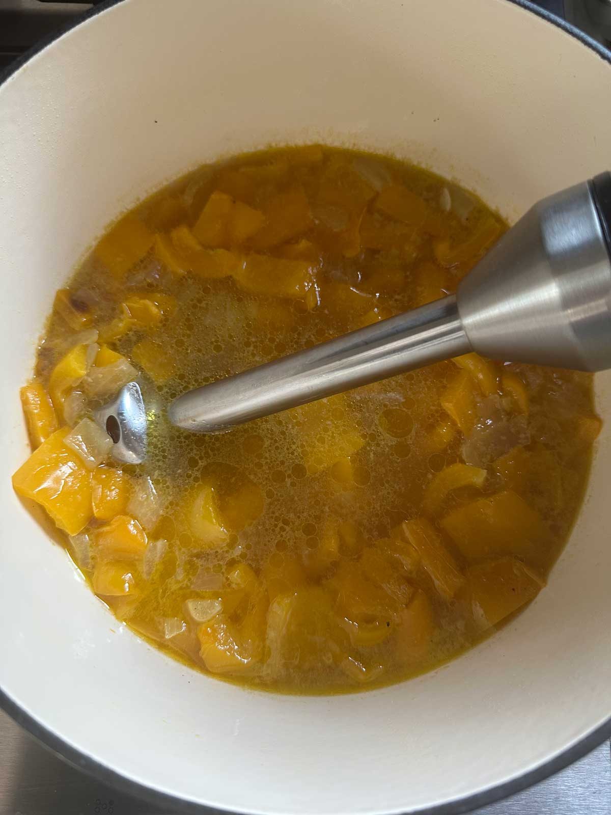 A pot containing chopped yellow vegetables and broth, with an immersion blender partially submerged, ready to blend the mixture into a soup or puree.
