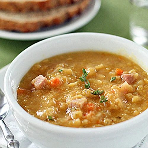 A bowl of thick split pea soup with diced ham and vegetables, garnished with fresh thyme. A spoon rests beside the bowl, with slices of whole-grain bread and a glass of water in the background.