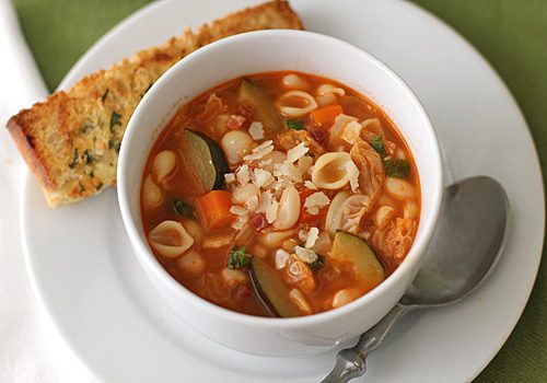 A bowl of vegetable soup with pasta, zucchini, beans, and carrots, topped with grated cheese. The bowl is on a white plate with a metal spoon and a piece of garlic bread on the side.