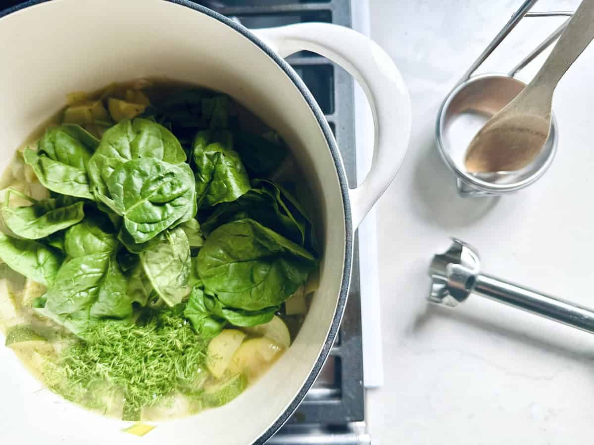 A pot on a stove contains fresh spinach, chopped vegetables, and herbs—ingredients for a Zucchini Soup with Dill and Greek Yogurt. Nearby are a wooden spoon in a metal holder and an immersion blender, suggesting comforting soup preparation.